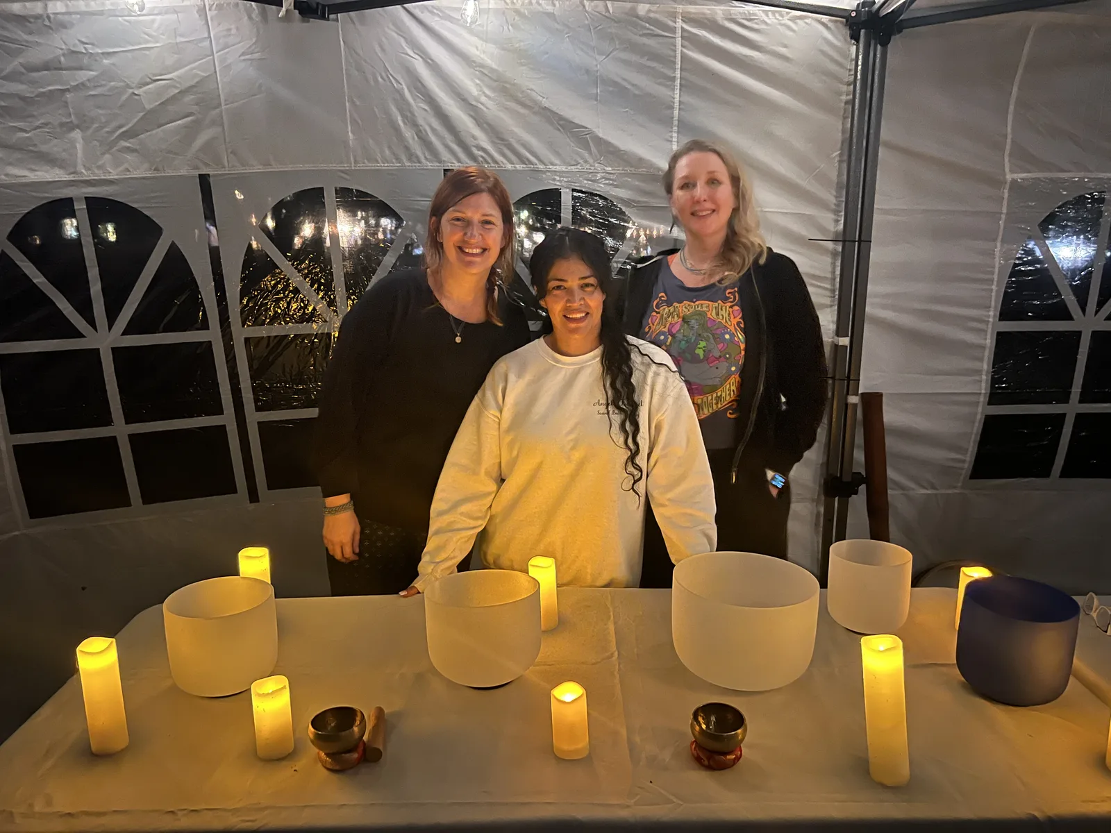 Three women smiling together behind a table of crystal singing bowls at the Becoming workshop